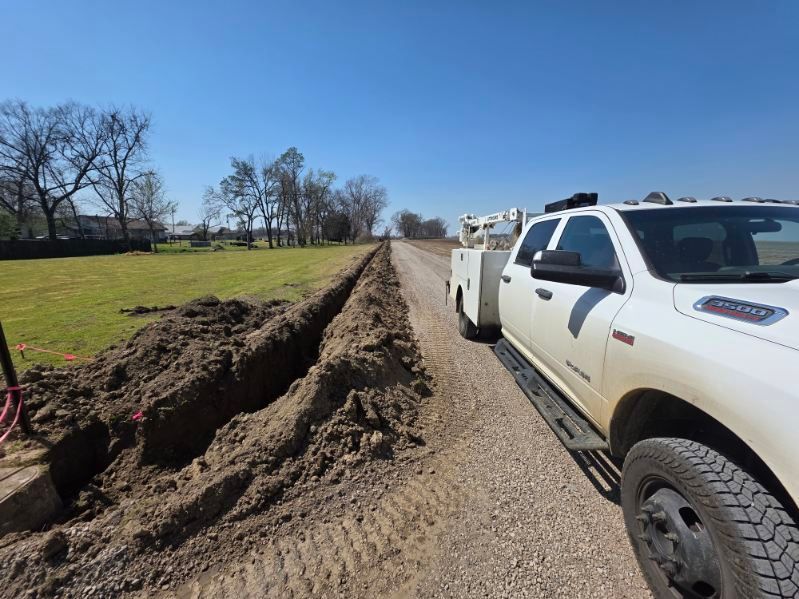 White pickup truck towing equipment on a dirt road beside a long trench in a grassy field