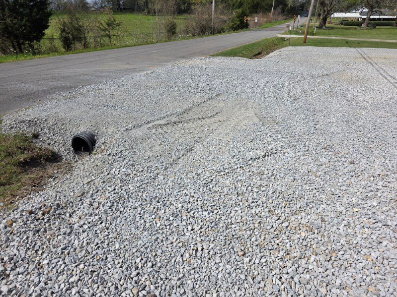 Gravel driveway with a drainage pipe and a washed-out ditch beside a paved road.