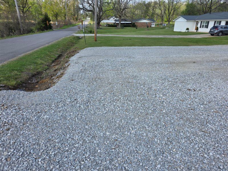 Gravel driveway beside a quiet road and grassy yard with houses in the background.