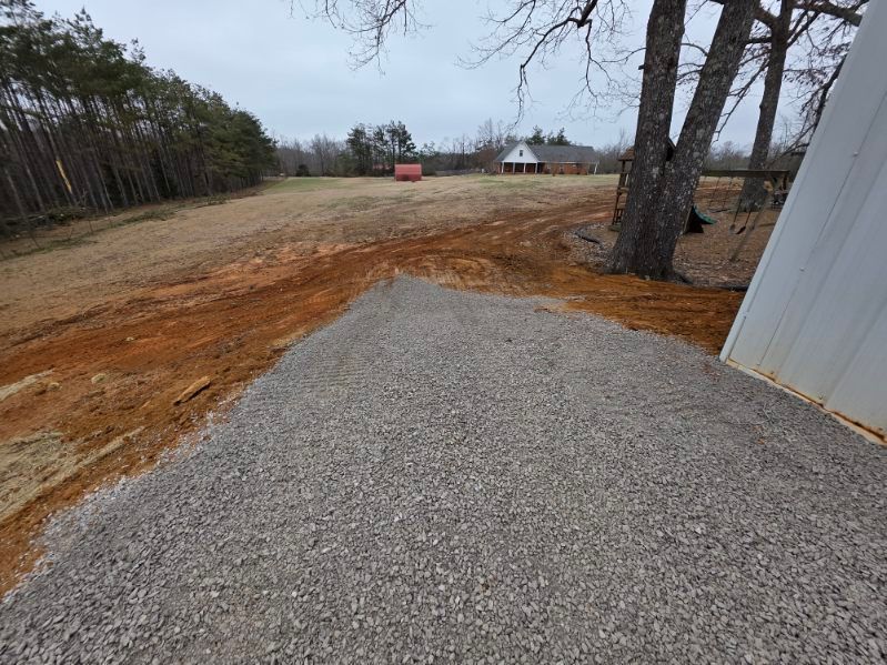 Gravel pile in front of a dirt yard, with a treeline and house in the background on an overcast day.