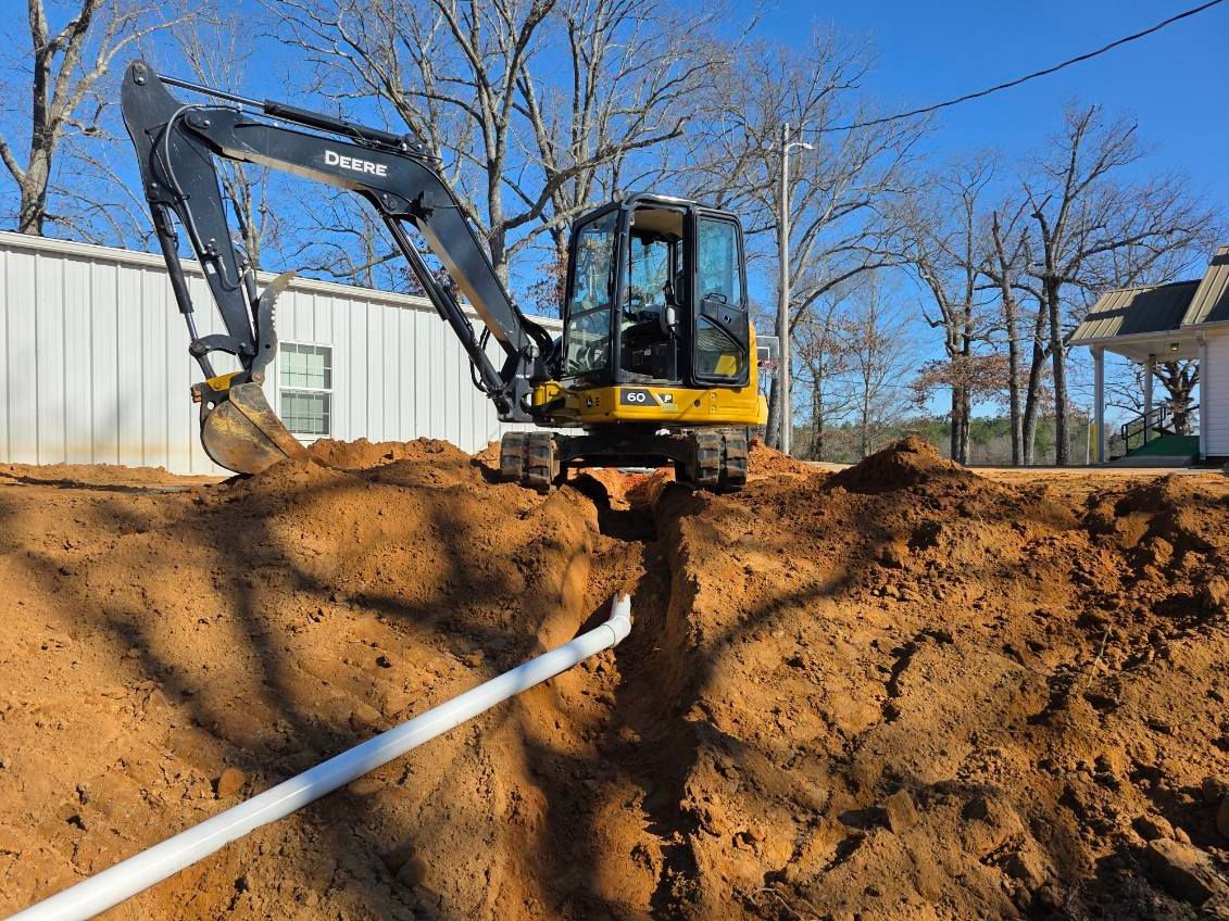 Yellow excavator digging trench; white pipe visible; sunny day.