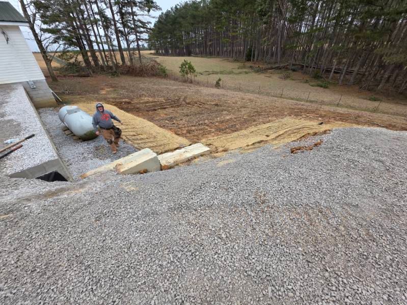 Person spreading mulch beside a house, with gravel driveway and wooded yard in the background