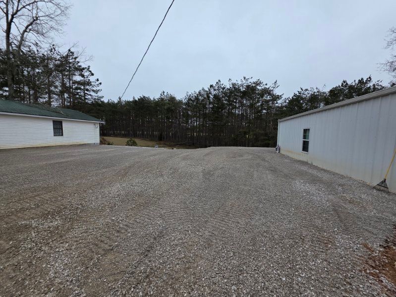 Gravel driveway between two buildings, white and light green, with treeline in the background under an overcast sky.