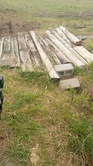 Stacked weathered wooden beams beside cinder blocks in a grassy field.