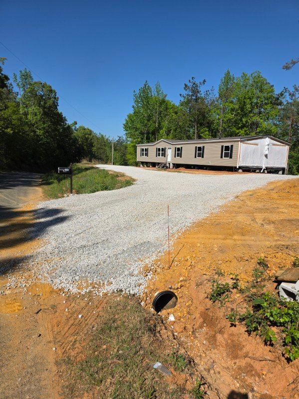 Gravel driveway leading to a beige mobile home in a wooded clearing under a clear blue sky