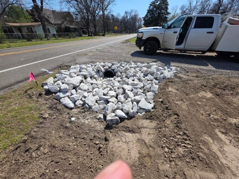 White rocks around a drainage basin beside a dirt shoulder, with a white pickup truck parked nearby.