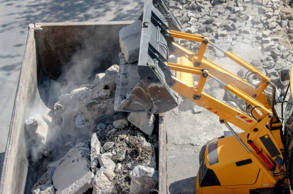 Yellow excavator loading rubble into a gray container, creating dust.