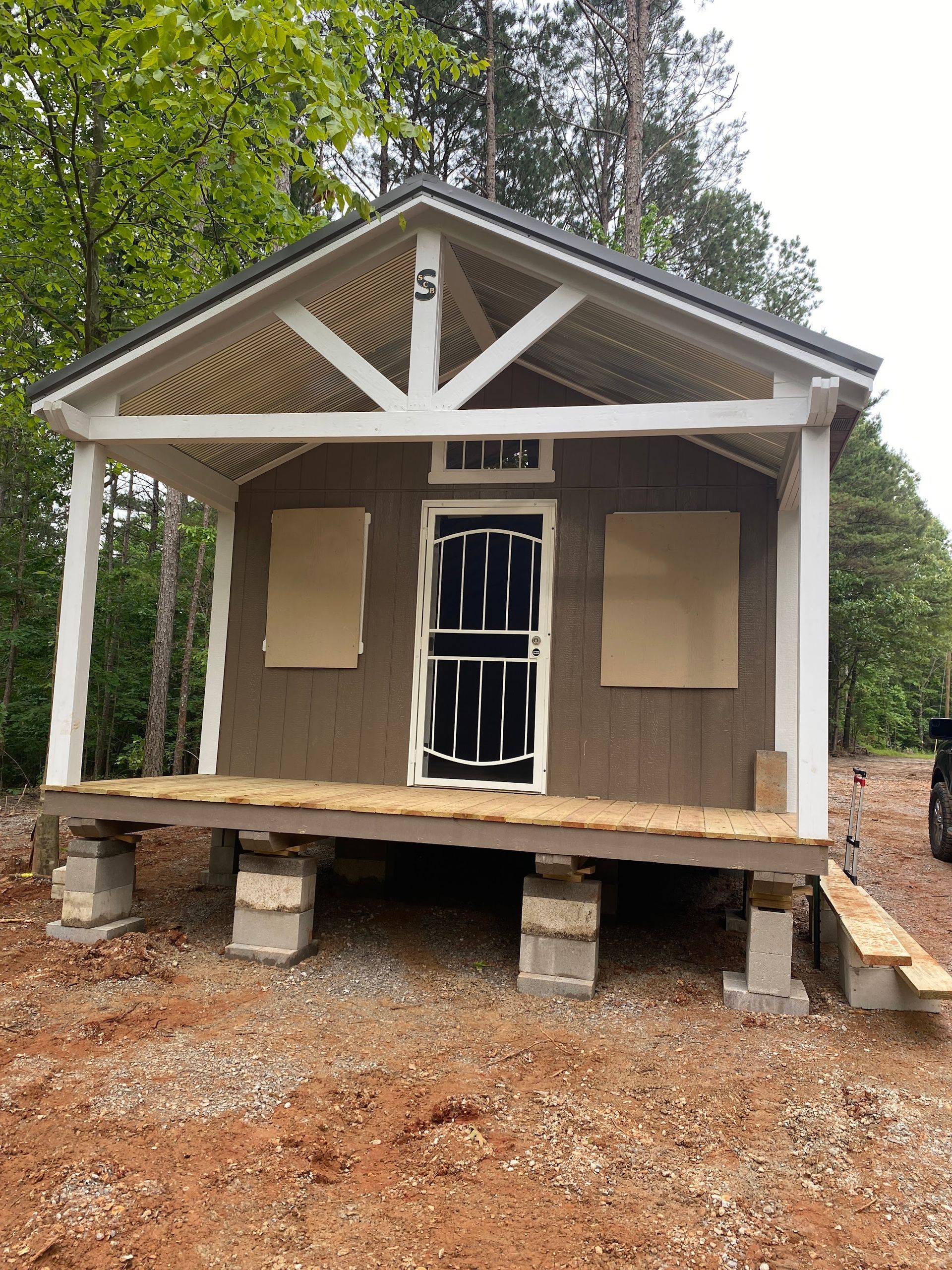 Small brown cabin with porch, white trim, and cinder block foundation, in a wooded area.