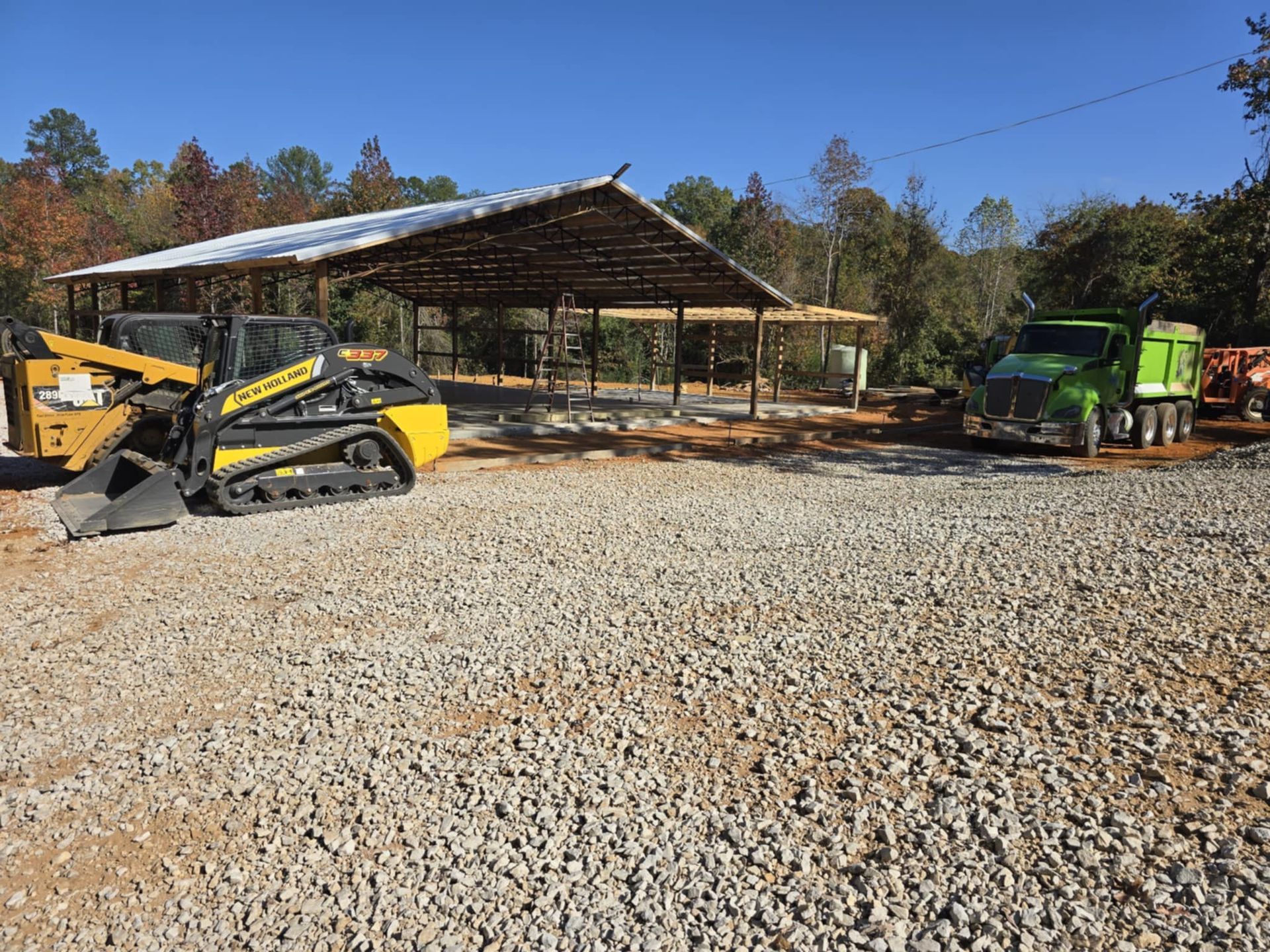Yellow skid steer and green dump truck on gravel lot in front of wooden shelter under a blue sky.