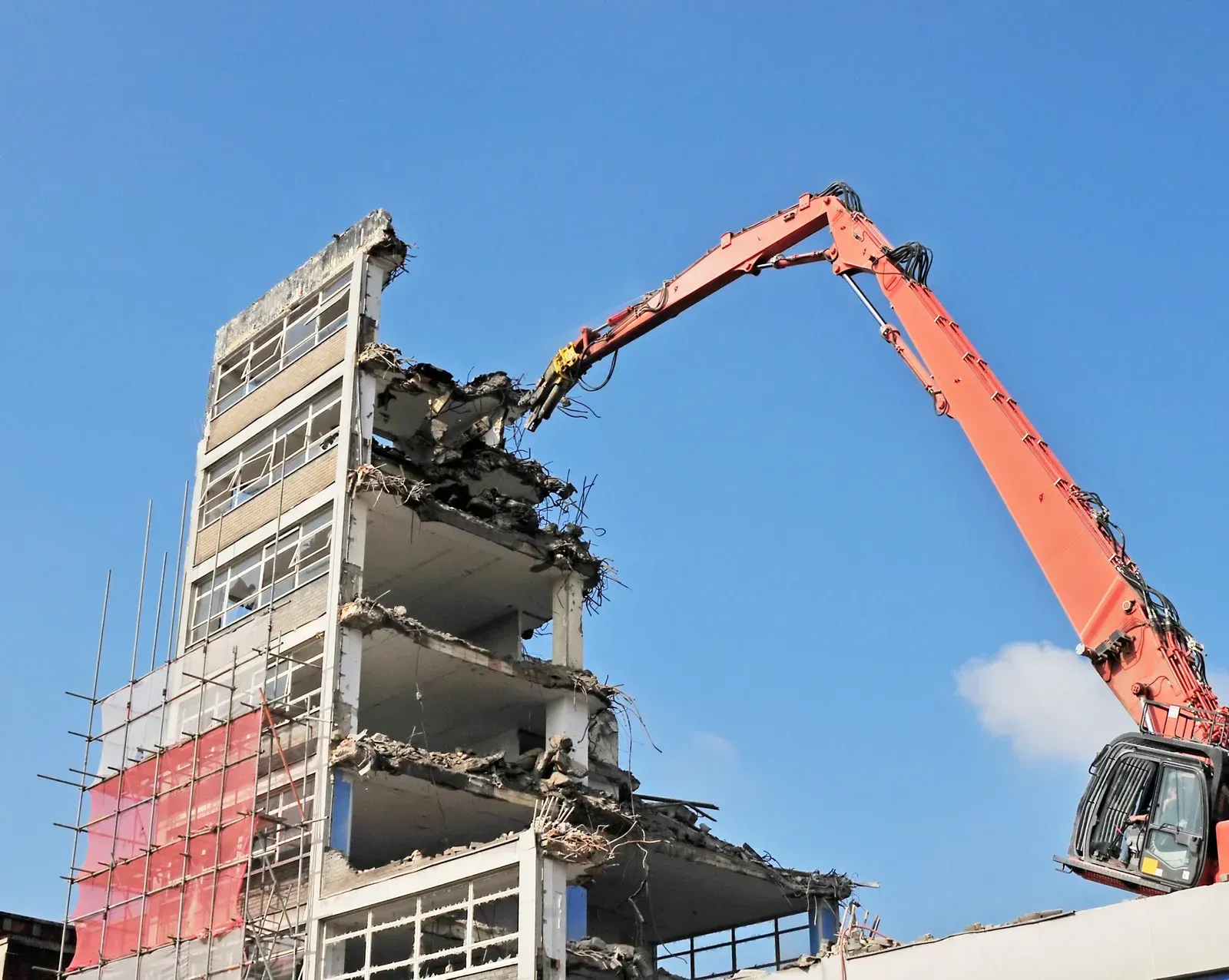 A large excavator demolishes a multi-story building under a blue sky.