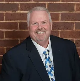 A man in a suit and tie is smiling in front of a brick wall.