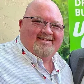 A bald man with glasses and a beard is smiling in front of a green sign.