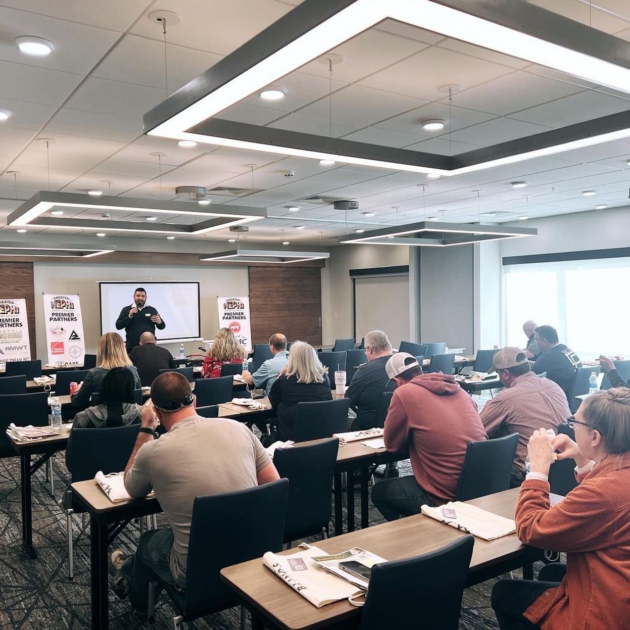 A man is giving a presentation to a group of people sitting at tables in a large room.