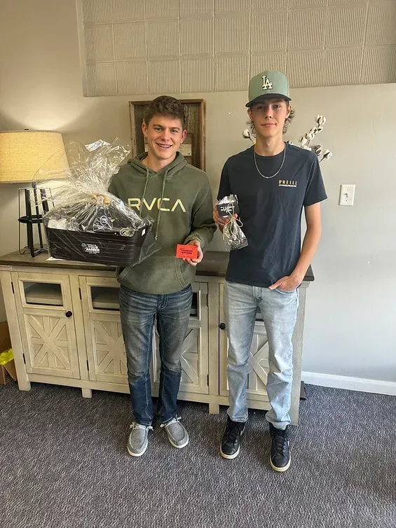 Two young men are standing next to each other in a living room holding trophies.