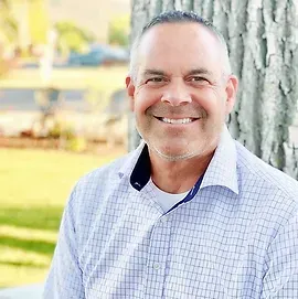 A man in a plaid shirt is smiling while leaning against a tree.