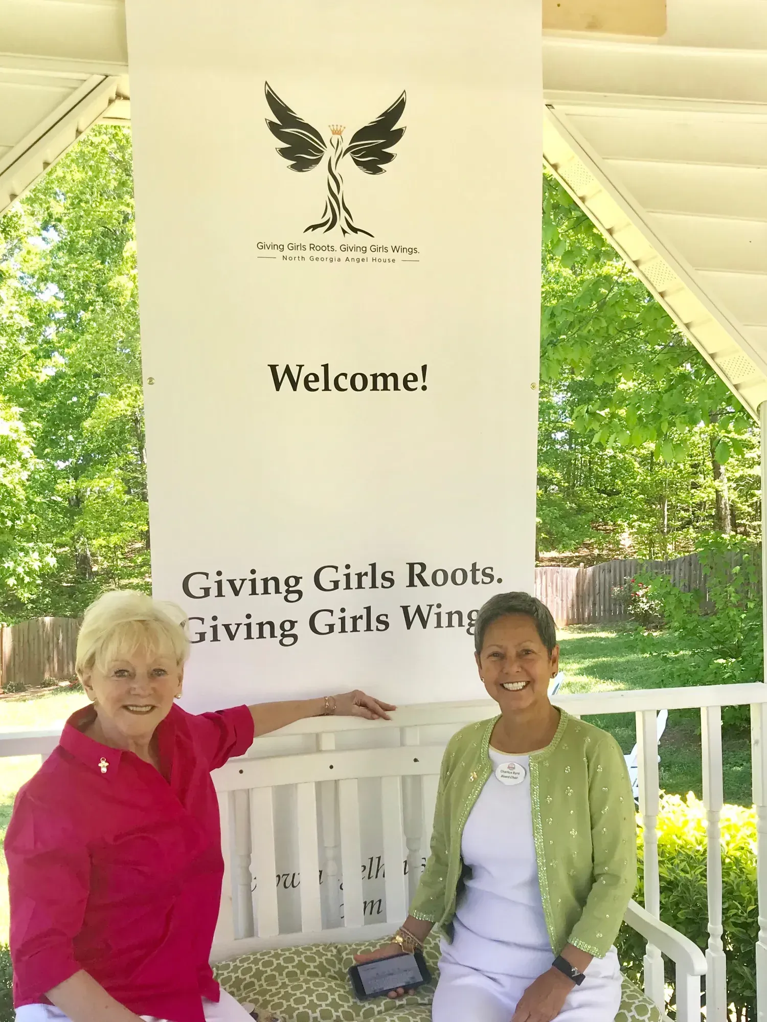Two women are sitting on a bench under a sign that says welcome giving girls roots giving girls wings