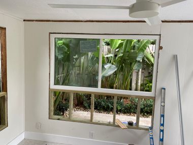 Interior view of a window being installed in a room with wooden framing, looking outside to lush greenery.