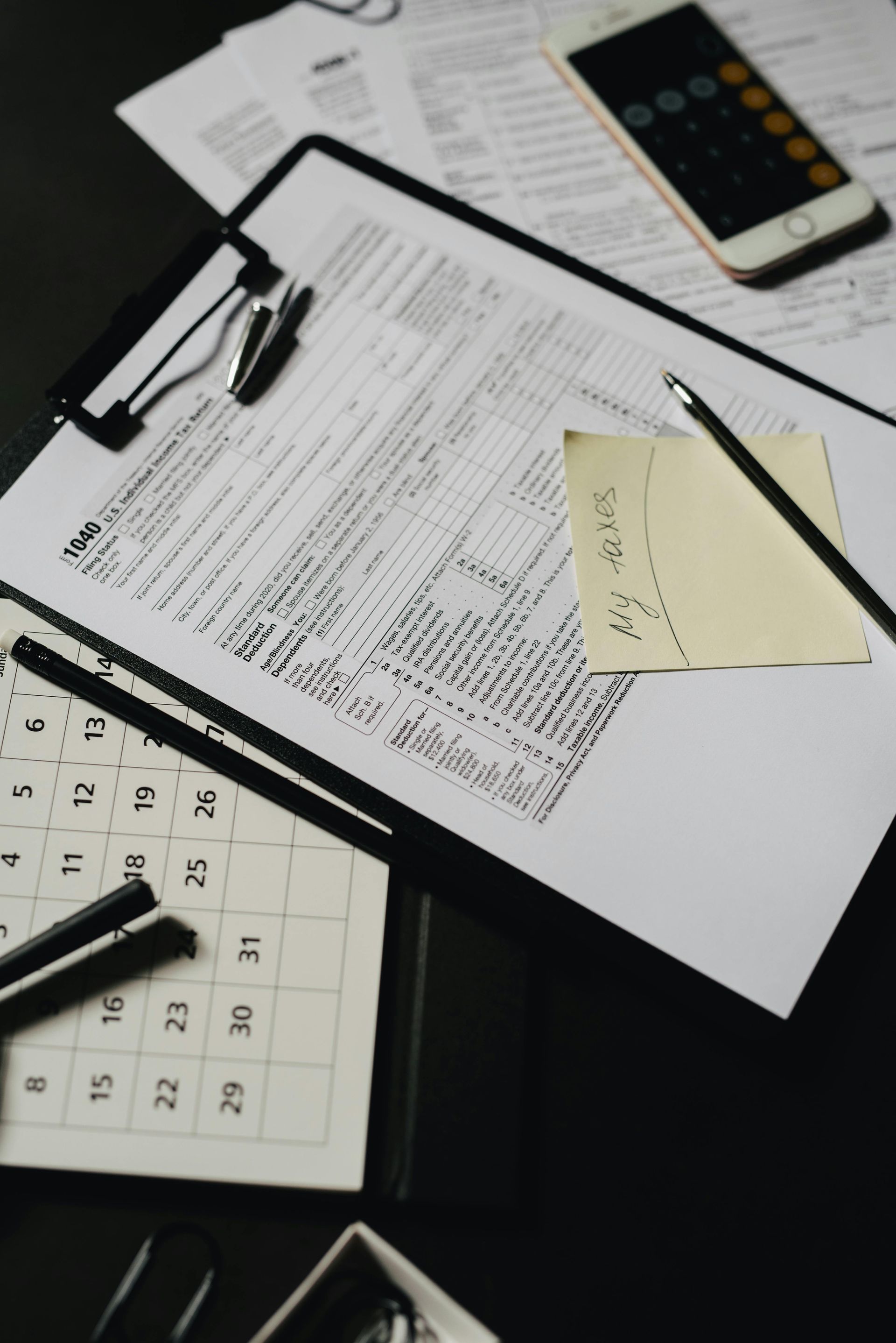 A desk with tax forms on a clipboard, a calendar, a calculator, pens, and a sticky note, representing financial planning.