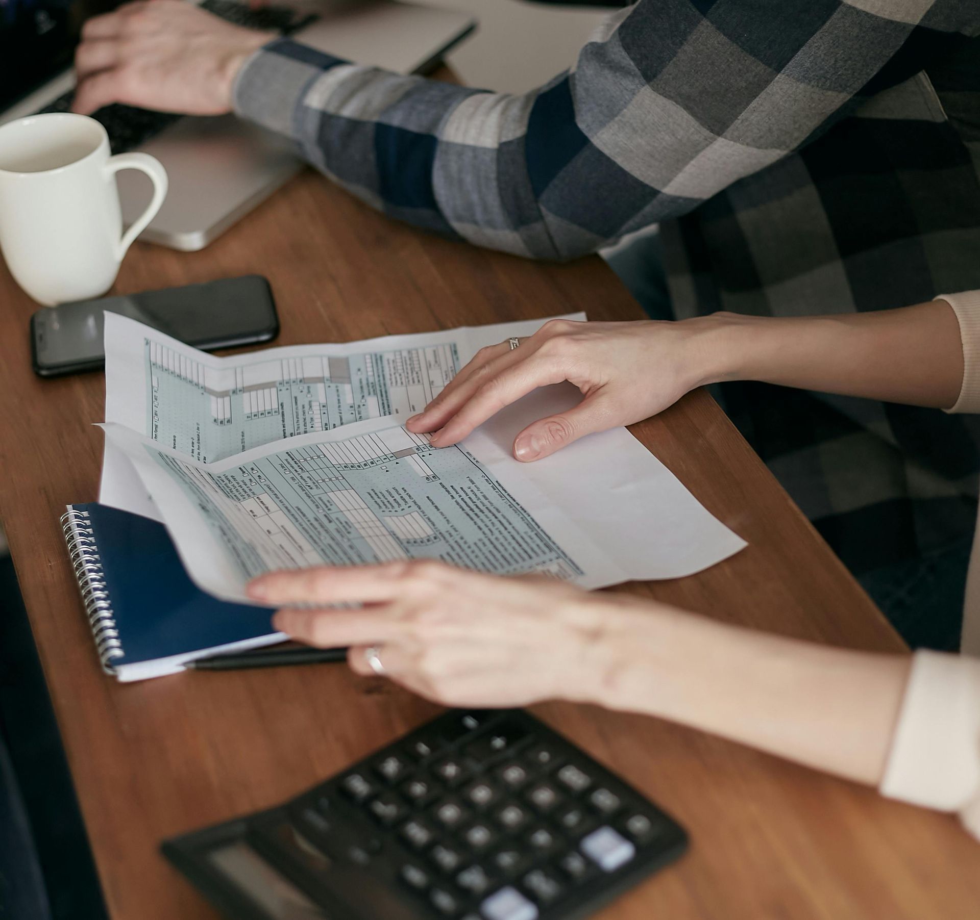 Two people sit at a wooden desk with a document, a calculator, a notebook, and a coffee mug, working on financial tasks.