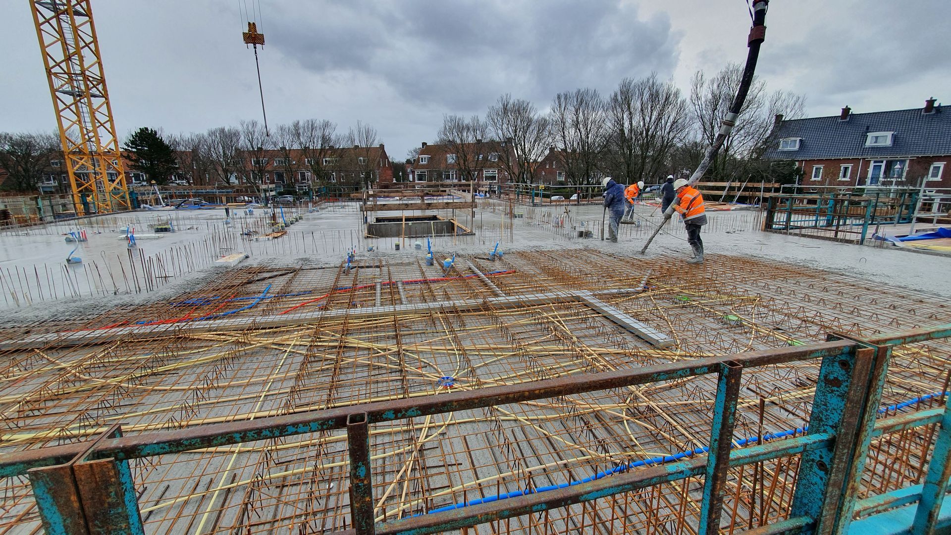 Bouwplaats met wapeningsstaal, arbeiders, kraan en gebouwen onder een bewolkte hemel.