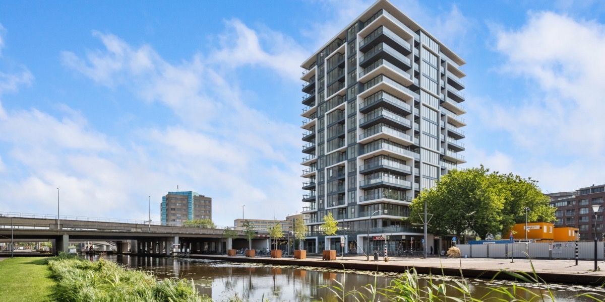 Een hoog, modern gebouw aan een kanaal in een stad, onder een helderblauwe hemel.