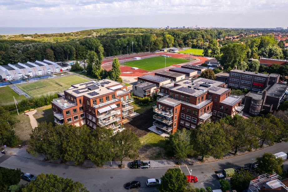 Luchtfoto van bakstenen appartementencomplexen, een sportveld en een groen landschap onder een blauwe hemel.