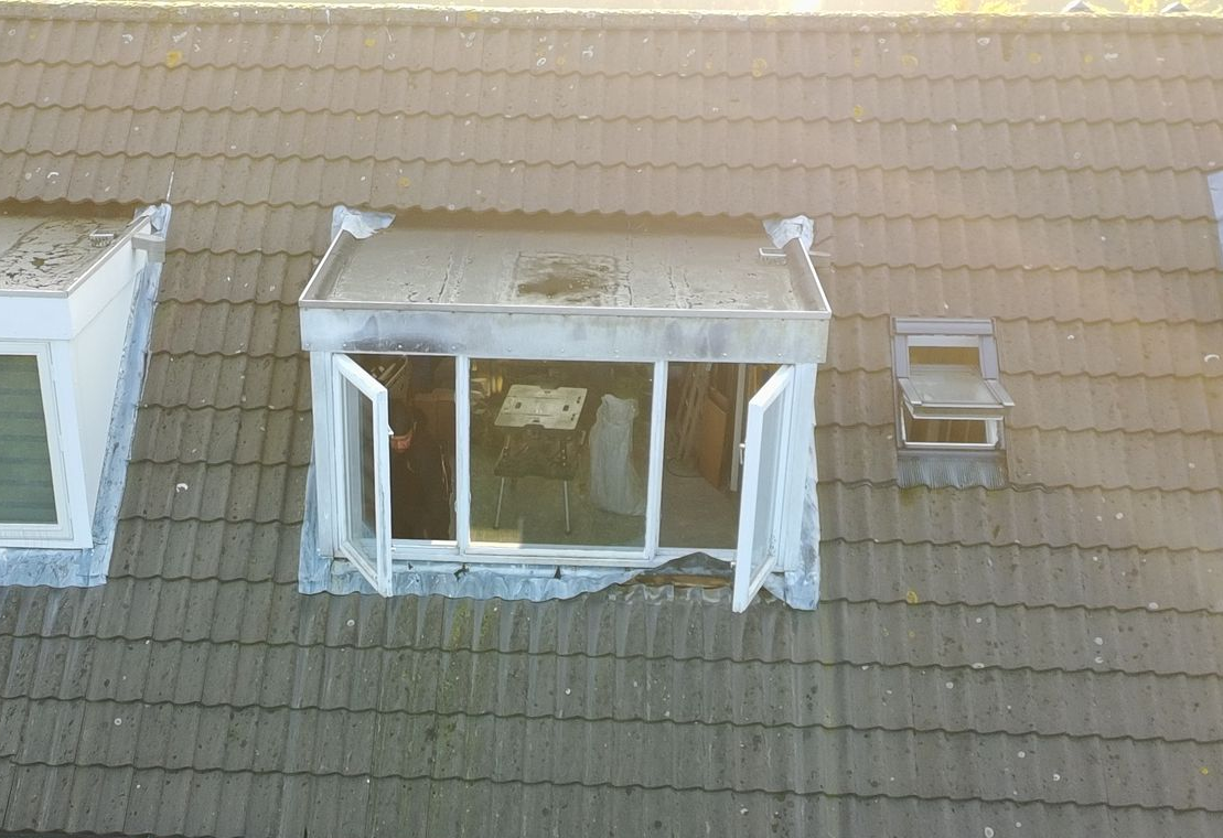 Dak met open raam. Grijze dakpannen, verweerde raamkozijnen. Zonnige buitenverlichting.