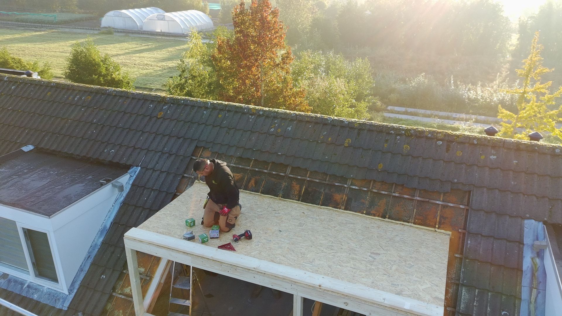Een dakwerker werkt aan een gedeelte van een plat dak, omringd door gereedschap, met een veld en bomen op de achtergrond.