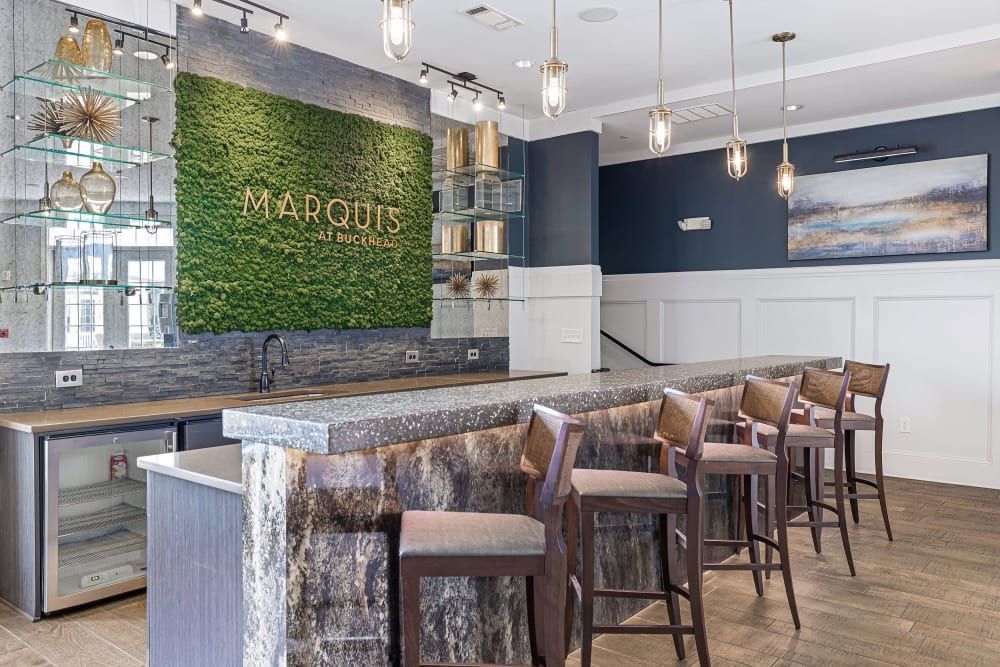 Modern bar area in a stylish apartment complex with high stools at a polished stone countertop, a green moss wall with gold lettering 'MARQUIS at Buckhead', elegant hanging light fixtures, and a serene painting on the wall at Marquis at Buckhead in Atlanta, GA.