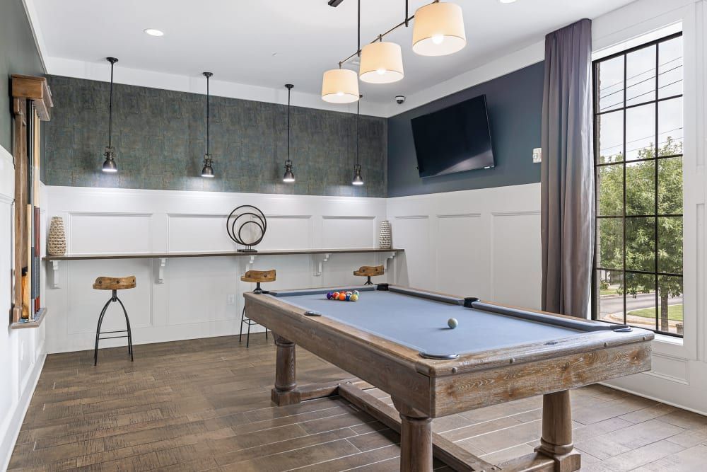 Modern game room interior with a blue pool table in the foreground, elegant pendant lights above, and a mounted flat-screen TV on a dark accent wall. A large window with curtains offers natural light, complementing the white paneling and decorative wall art at Marquis at Buckhead in Atlanta, GA.