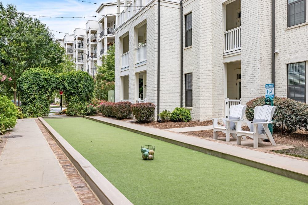 A well-maintained outdoor common area at an apartment complex featuring a narrow strip of artificial green turf with a bocce ball court setup, flanked by a concrete walking path. White Adirondack chairs sit nearby on a small resting area next to a building with white brick exterior and multiple balconies. String lights are hung overhead and lush greenery with blooming flowers adorn the space, creating an inviting urban living environment at Marquis at Buckhead in Atlanta, GA.