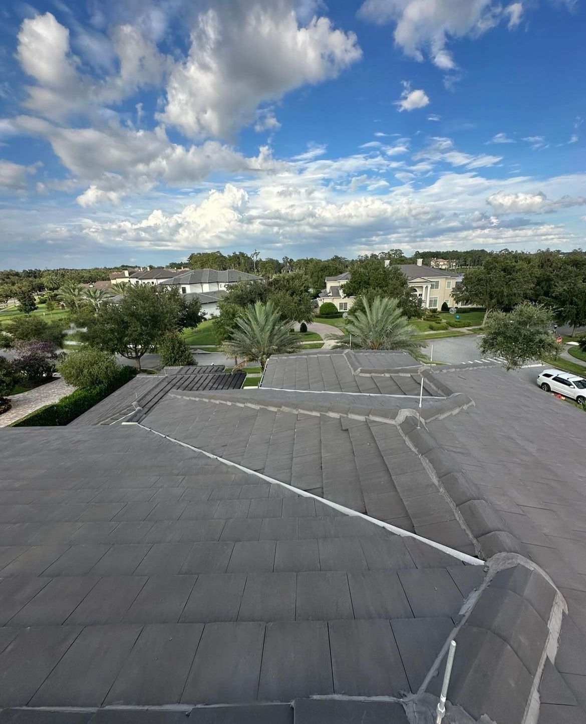 View from a dark roof overlooking a neighborhood with green trees, white houses, and a blue sky with clouds.