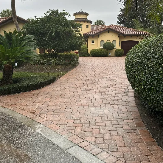 Brick driveway leading to a yellow house with a round tower, surrounded by green bushes and plants.