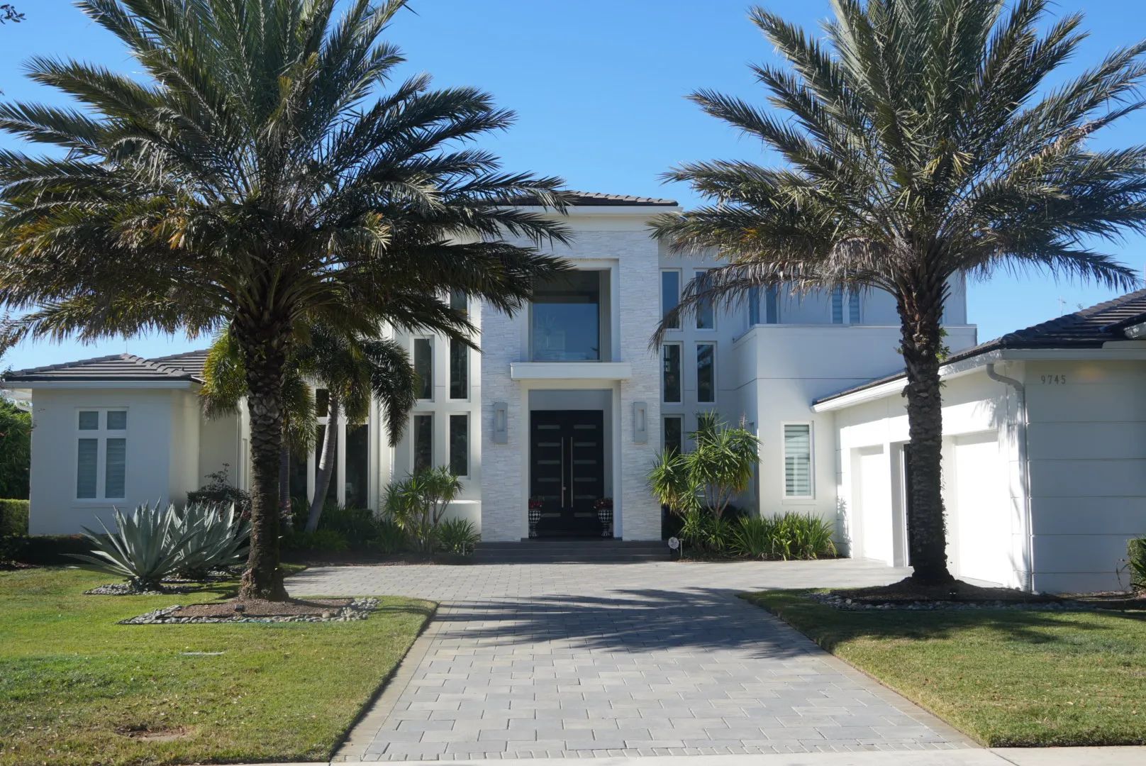 White two-story house with palm trees in front. Driveway leads to black front doors.