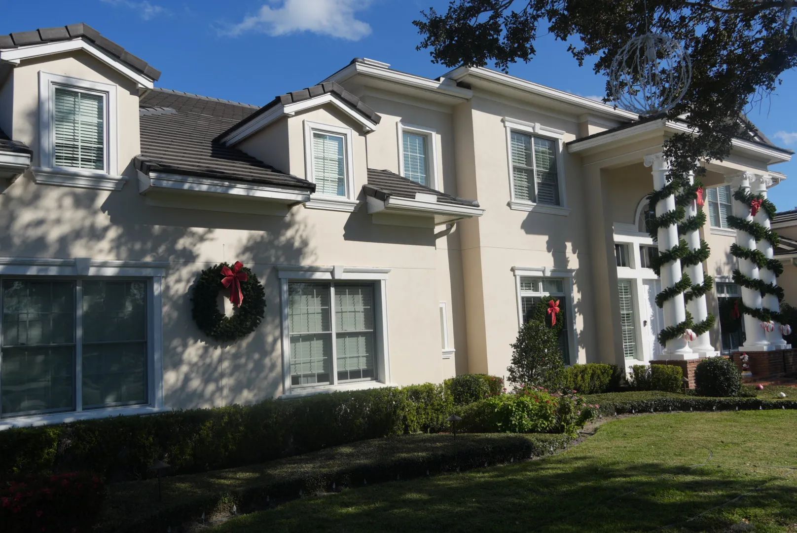 Beige two-story house decorated with Christmas wreaths and garland under a blue sky.