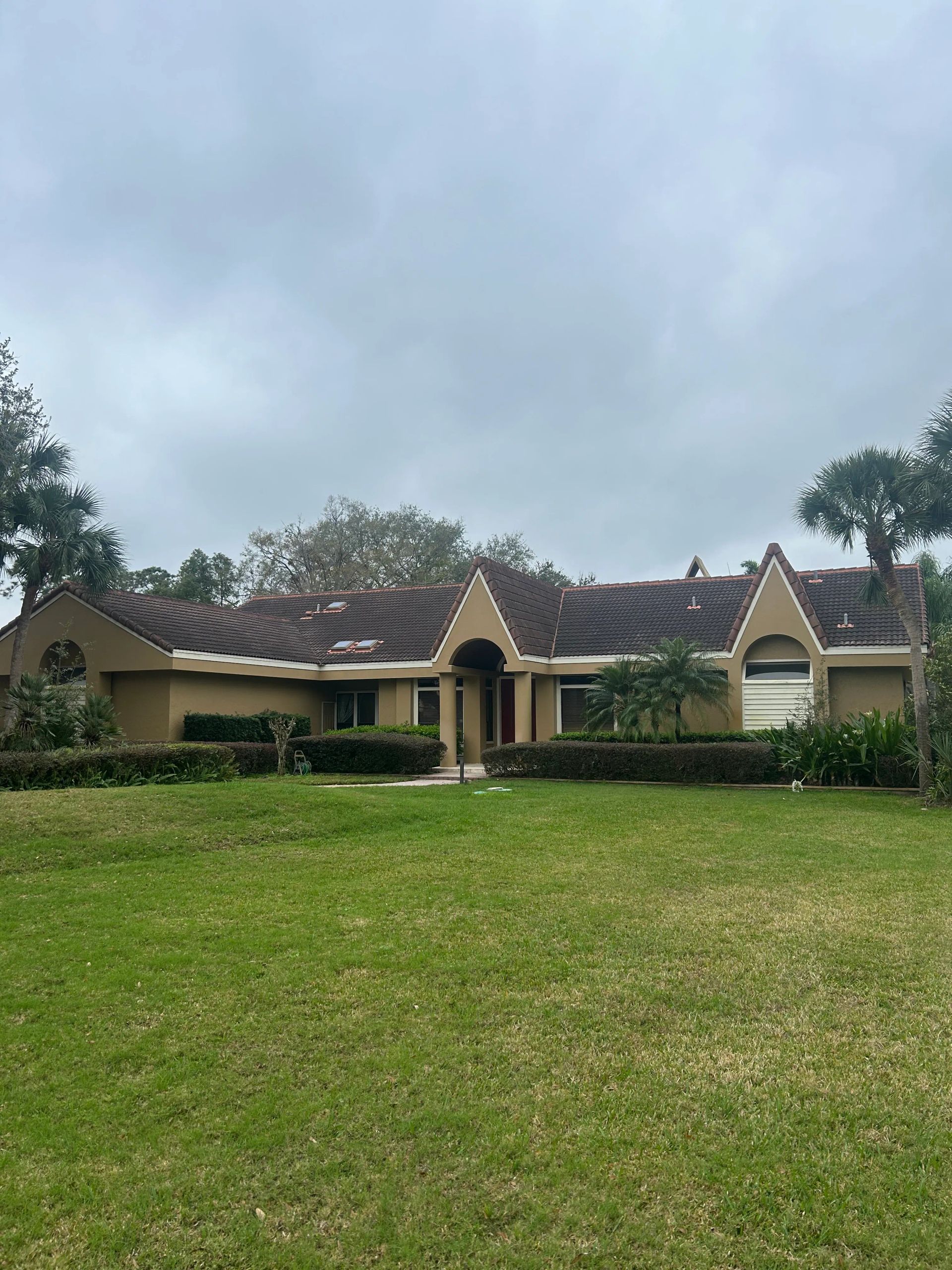 Tan ranch house with red tile roof, green lawn, and cloudy sky.