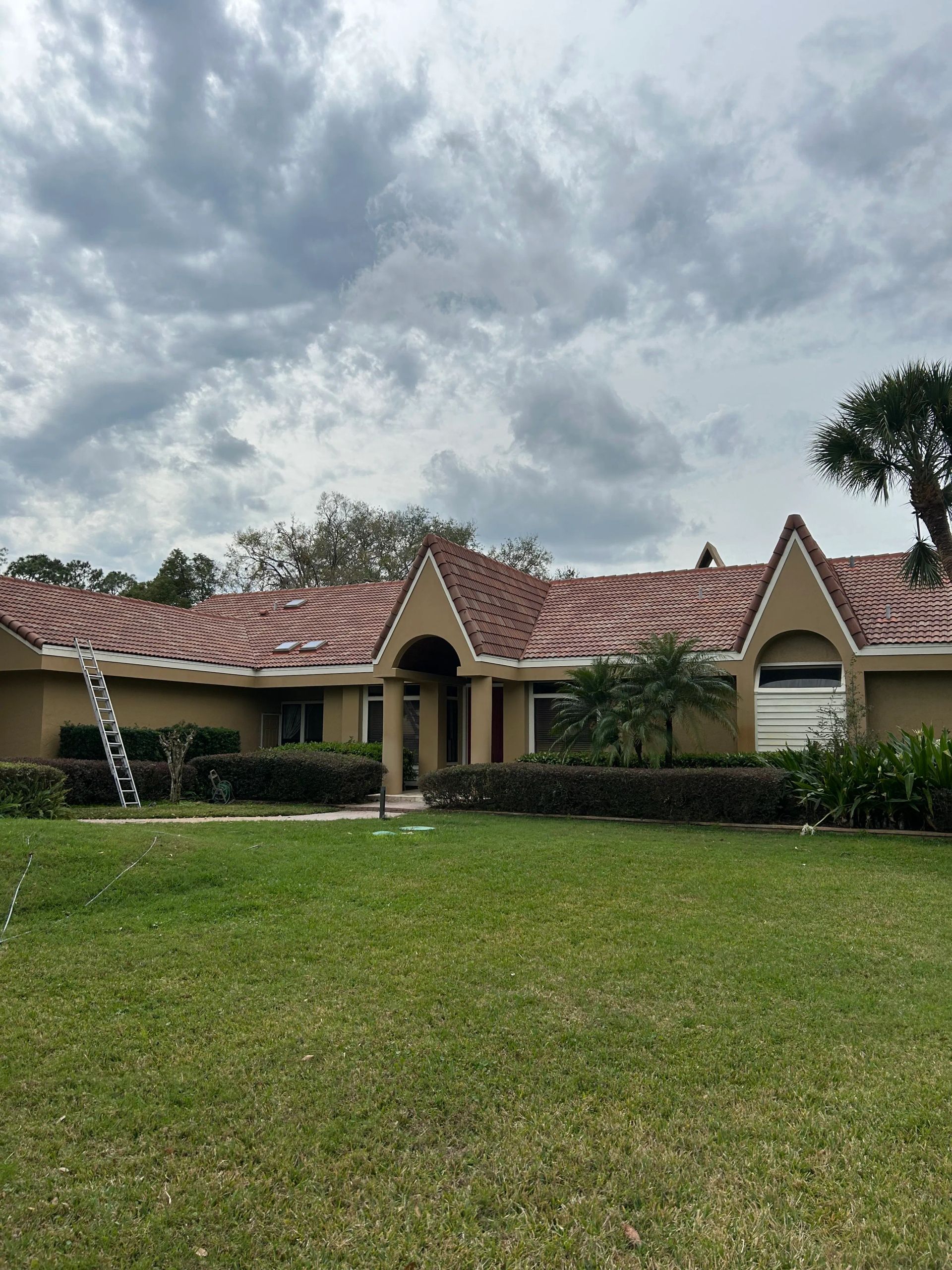 Tan house with red-tiled roof, green lawn, and cloudy sky. Ladder leaning on the side of the house.