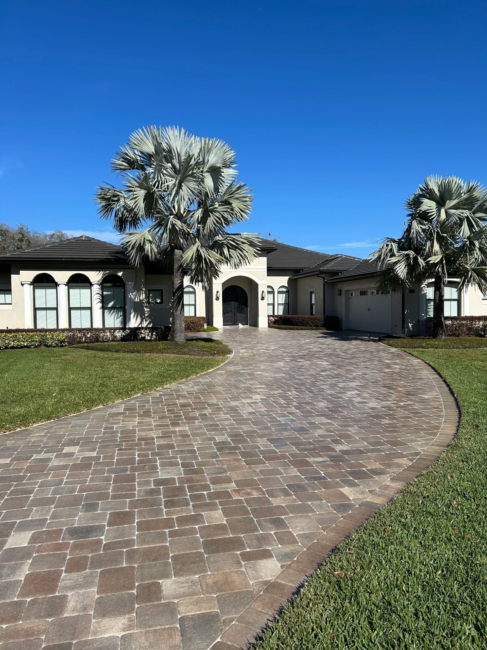 Elegant beige home with a brick driveway, framed by green grass and blue sky.