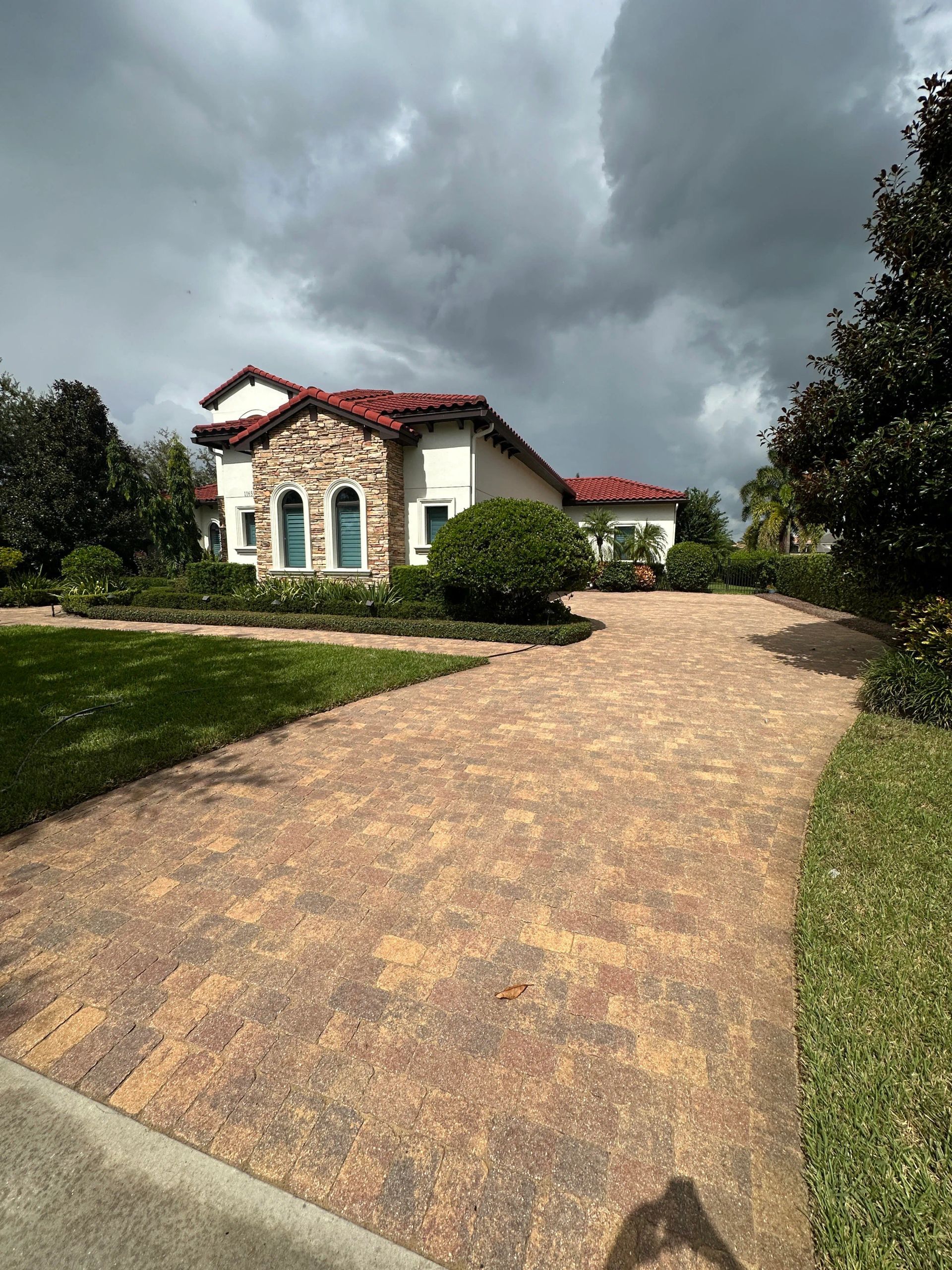A brick-paved driveway leads to a stucco house with a red-tile roof under a cloudy sky.