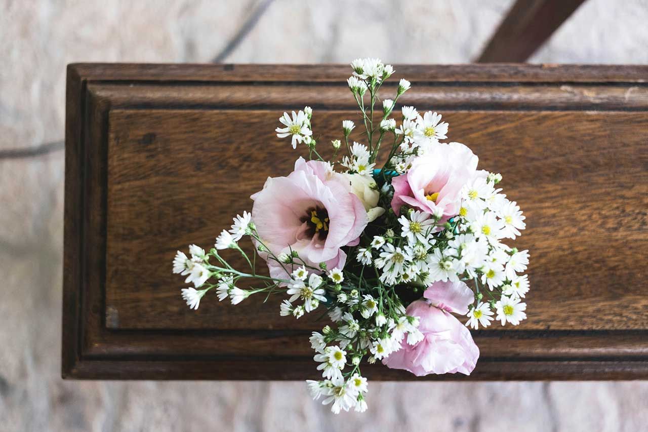 Small White And Pink Floral Arrangement Resting On A Wooden Casket During A Funeral Service