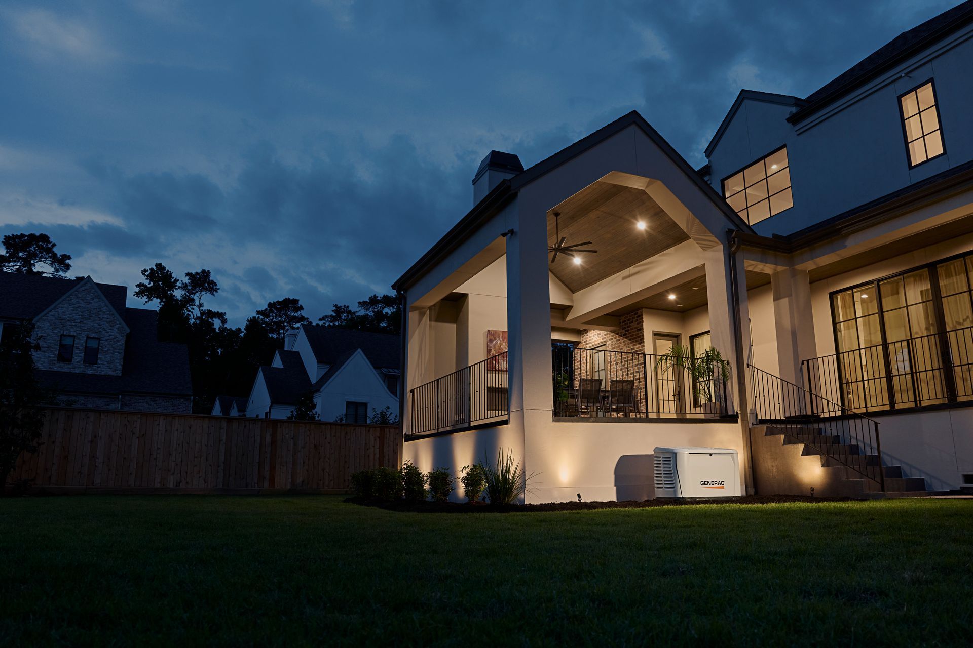 Back of a house at dusk with an illuminated porch, a grassy lawn, and a generator sitting near the exterior wall.
