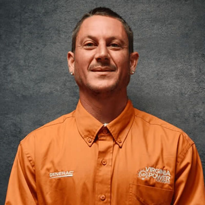Man in an orange Virginia Power shirt with a logo. He is smiling and wearing earrings, against a grey background.