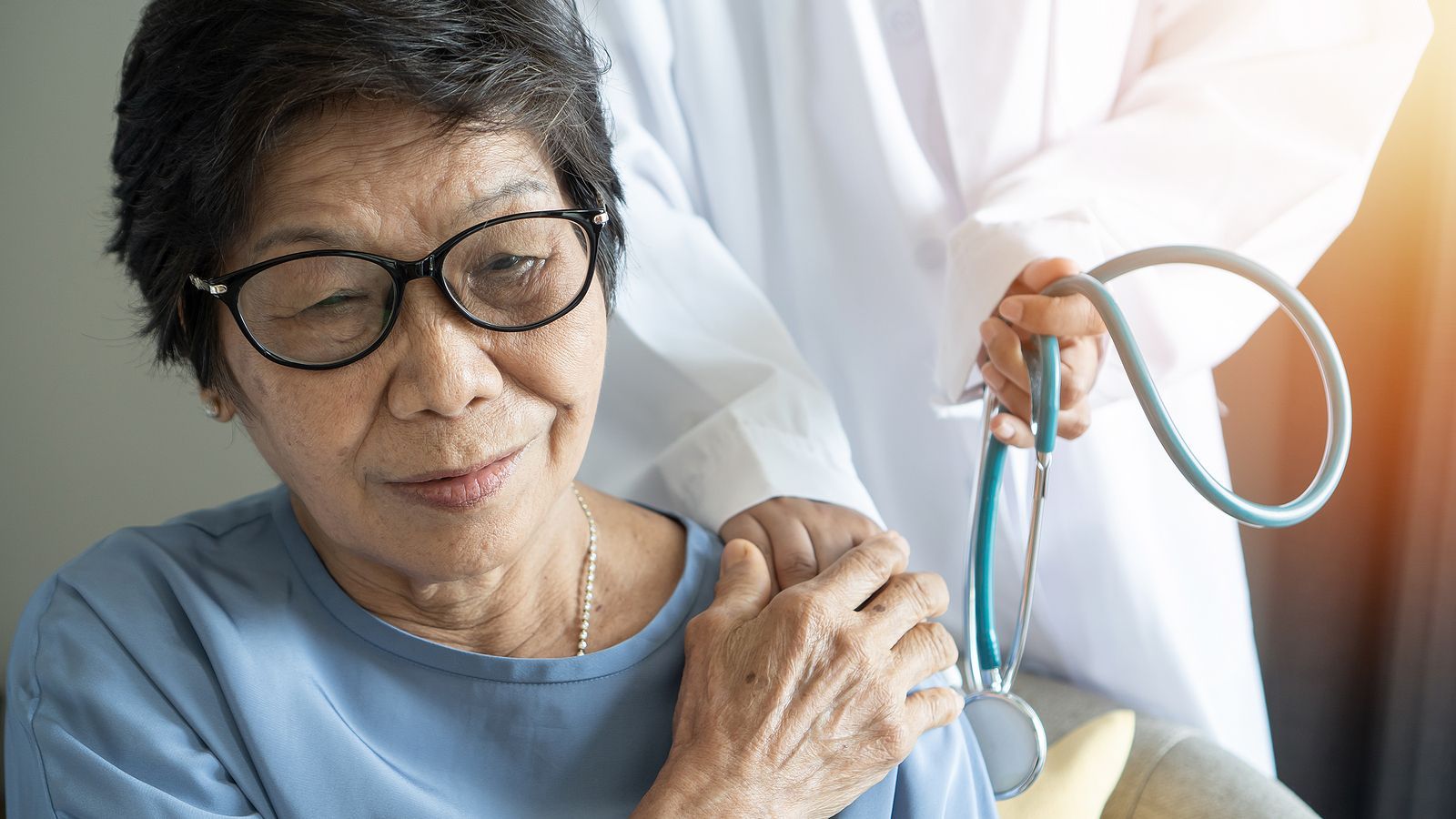 An older adult in glasses is supported by a doctor holding a stethoscope, with sunlight.