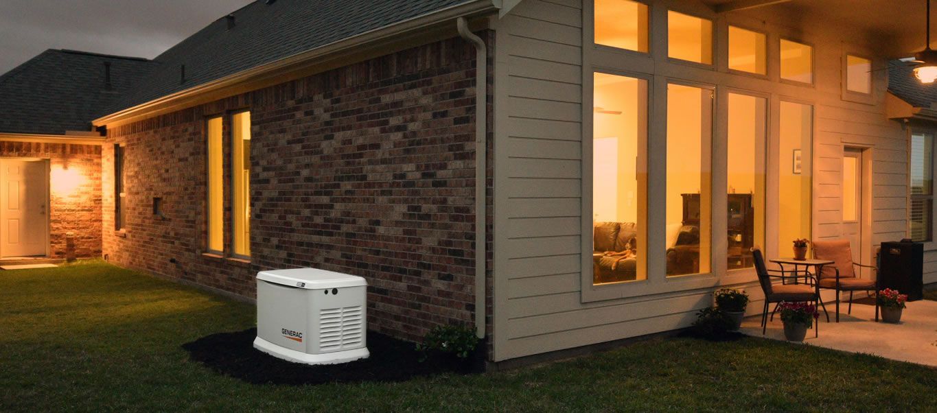 Backup generator next to a house with lights on at dusk. A patio with furniture is visible.