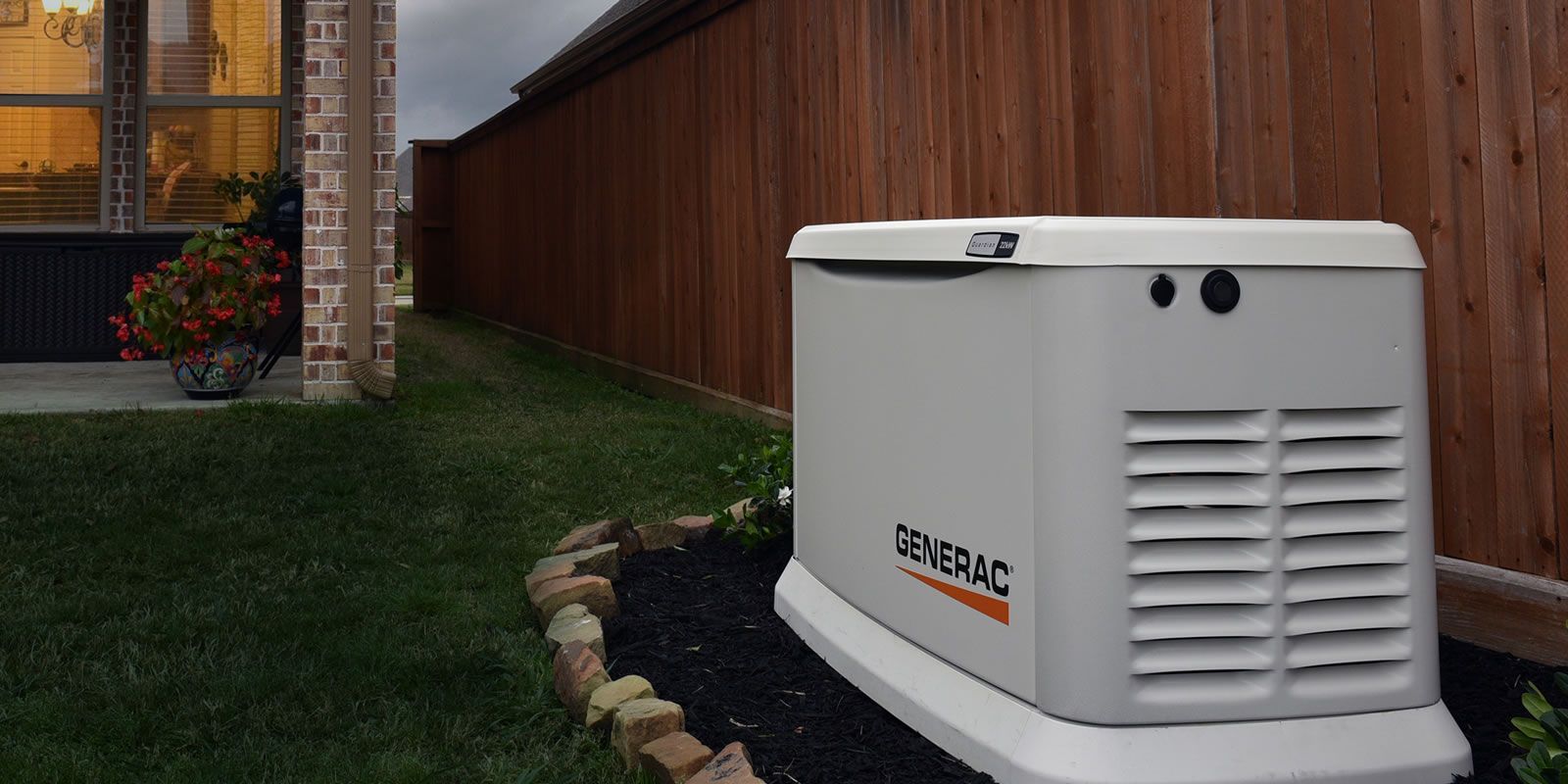 A Generac generator sits on a bed of black mulch in a backyard. A wooden fence and house are in the background.