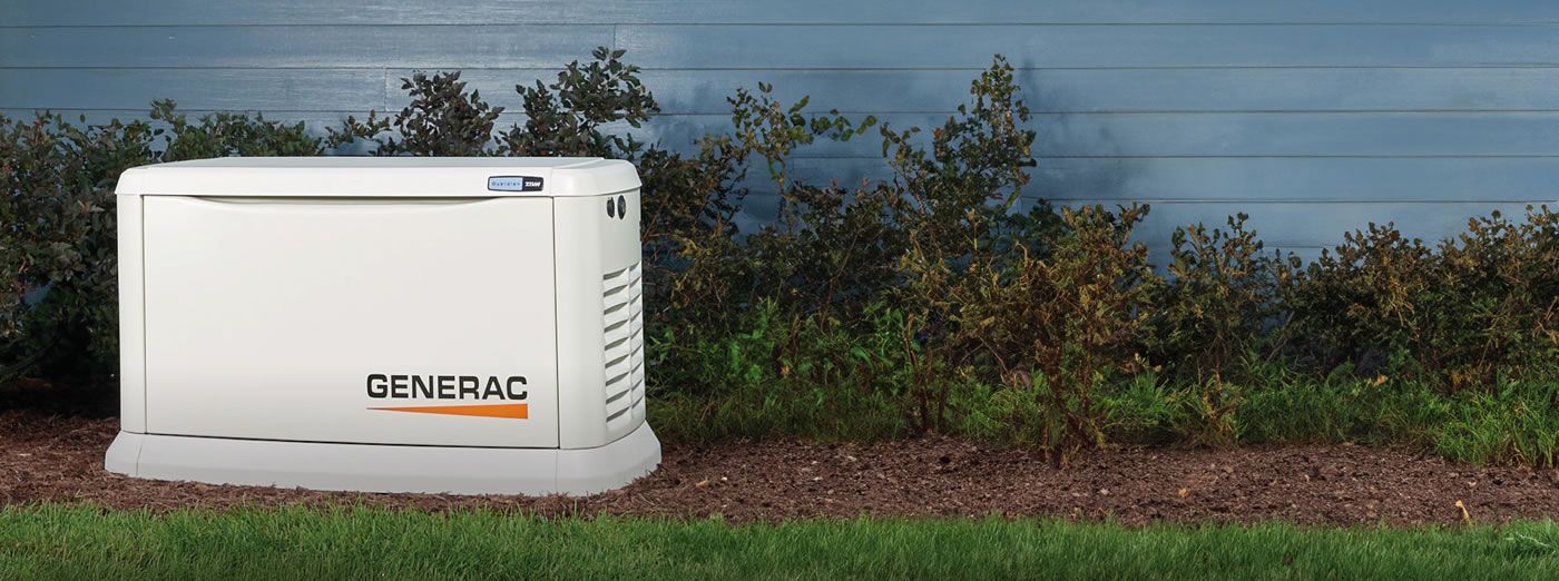 A Generac home generator next to a house with blue siding. Green grass and shrubs are in front of the generator.