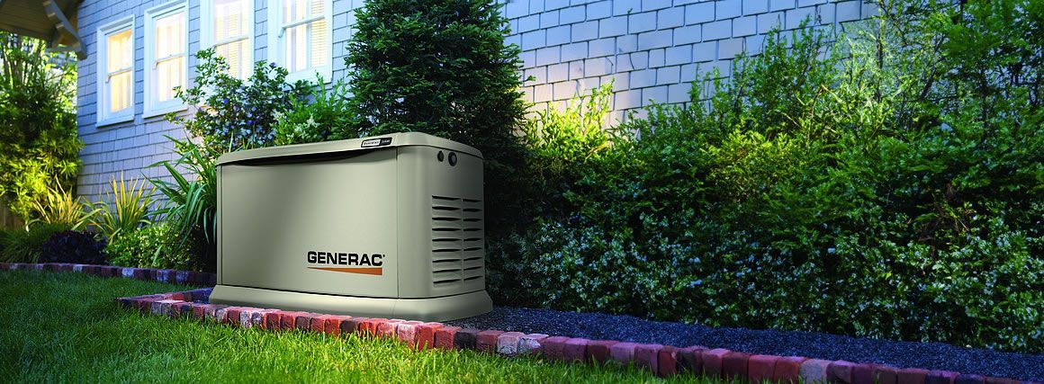 A standby generator is next to a house with illuminated windows and lush greenery.