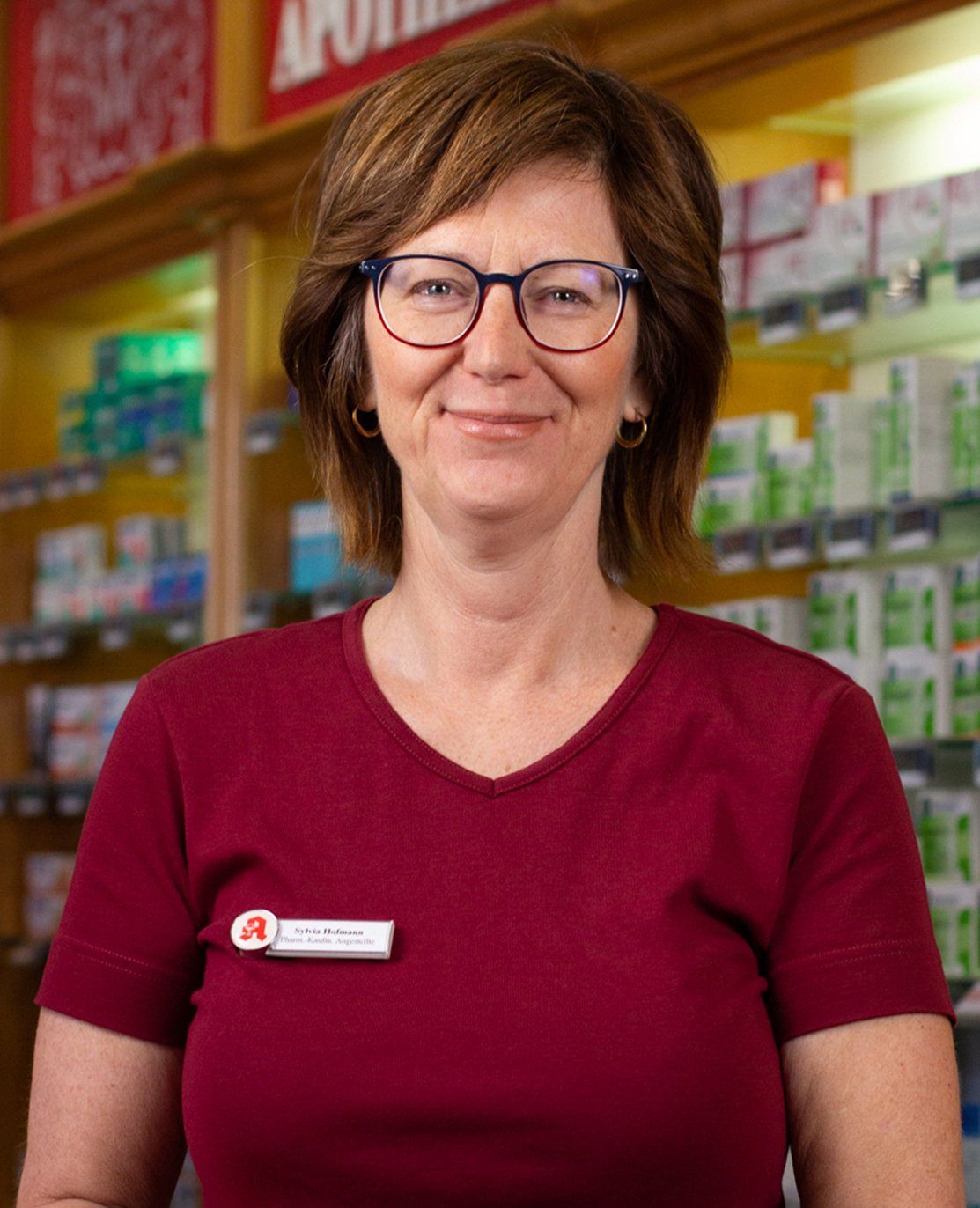 Eine Frau mit Brille und rotem Hemd steht vor einer Apotheke.