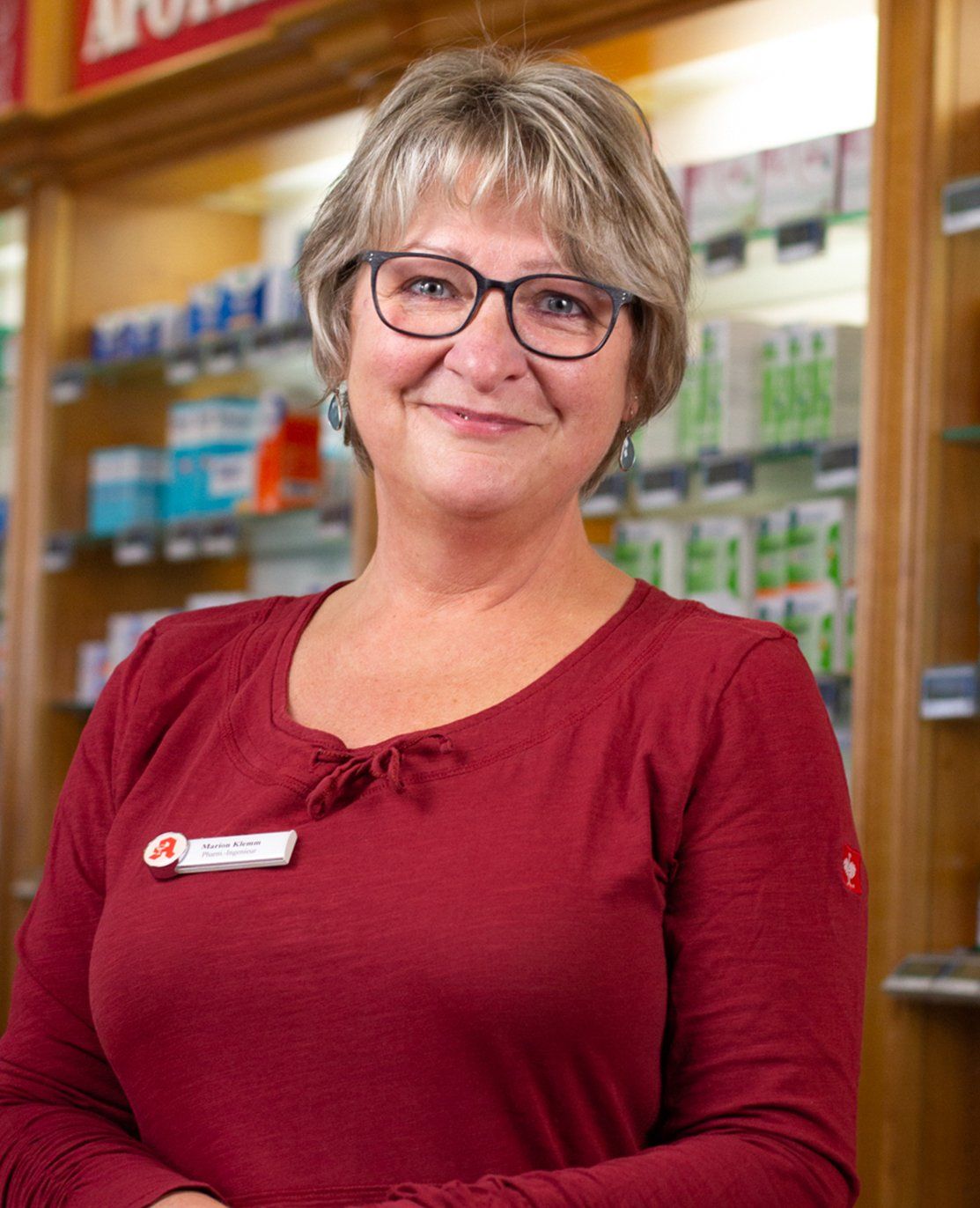 Eine Frau mit Brille und rotem Hemd steht vor einer Apotheke.