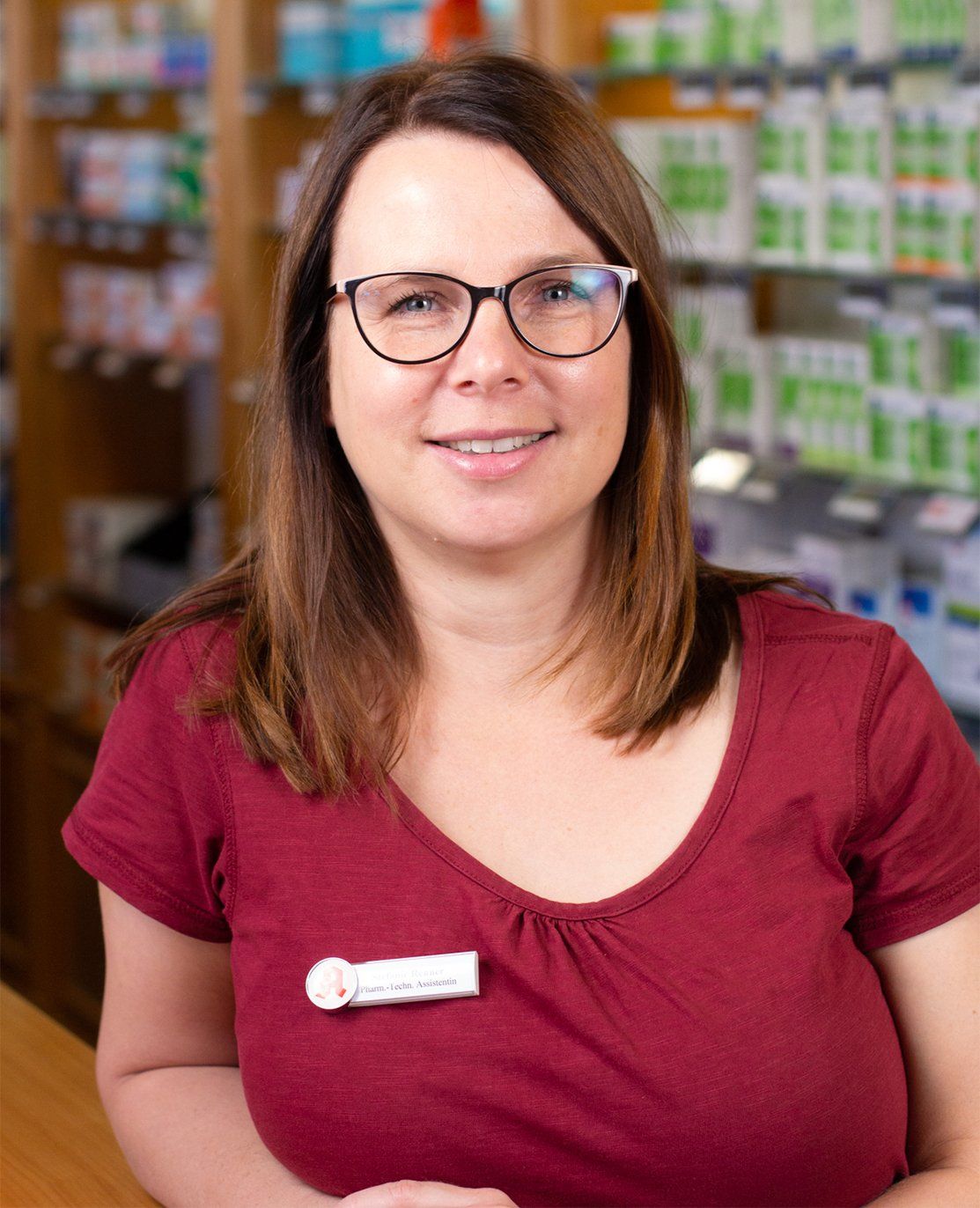 Eine Frau mit Brille und rotem Hemd steht vor dem Tresen einer Apotheke.
